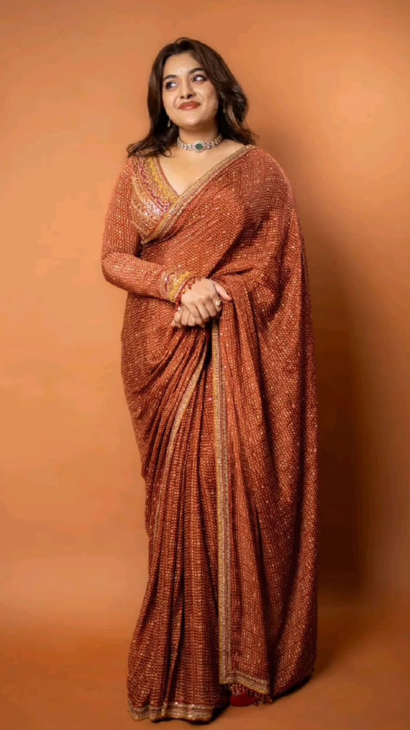 A woman dressed in a brown embellished saree with delicate shimmer detailing and a matching full-sleeve blouse. She styled the look with minimal jewelry and soft waves for a classy ethnic appearance.