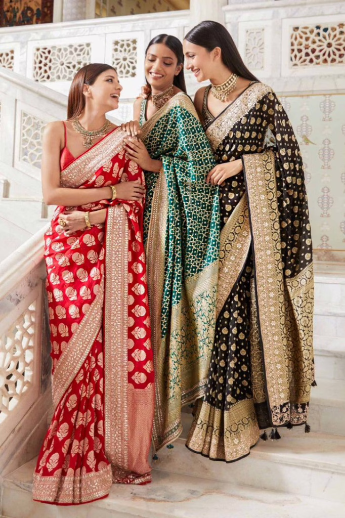 Three women wearing elegant Banarasi silk sarees in red, green, and black with intricate gold zari work, styled with traditional jewelry on a marble staircase.