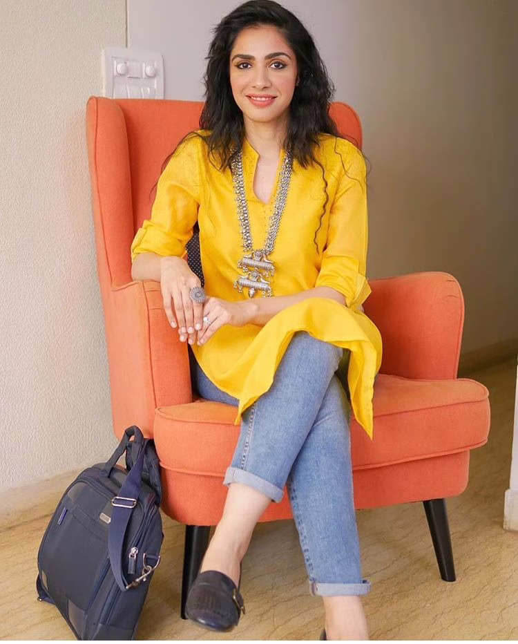 Woman wearing a bright yellow kurta with rolled-up denim jeans and statement silver jewelry, seated in a cozy indoor setting with a travel bag beside her.