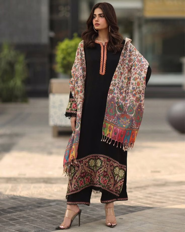 A woman wearing a black embroidered Kashmiri suit paired with a multicolored Kani weave shawl featuring intricate floral and paisley motifs with decorative tassels.