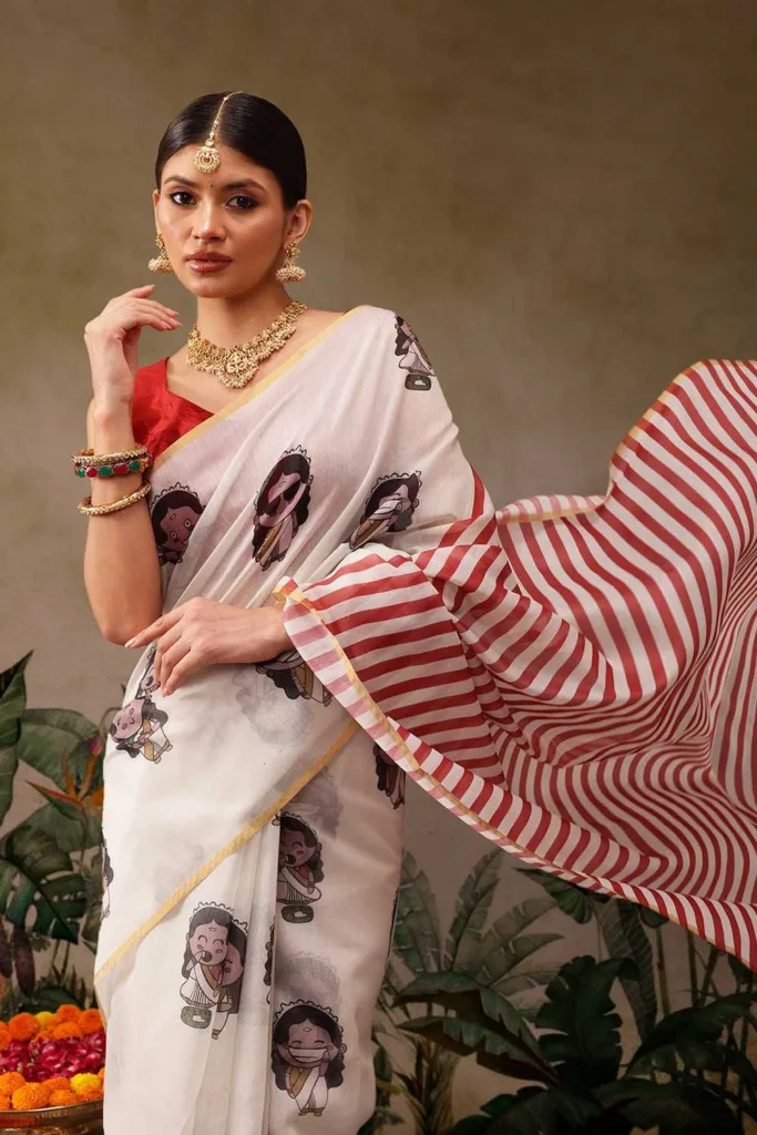 Woman wearing a white printed monsoon-friendly saree with cartoon motifs, a red striped pallu, and a matching red blouse. She pairs the saree with traditional gold jewelry, creating a stylish and comfortable look for festive celebrations, cultural events, and everyday ethnic wear.