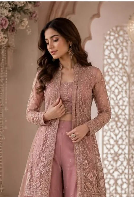 Dusty pink embroidered sharara suit featuring wide-leg floral embroidered sharara pants, a matching sleeveless blouse, and a heavily embellished long jacket with sheer detailing, paired with a flowing net dupatta in an elegant palace-inspired setting.