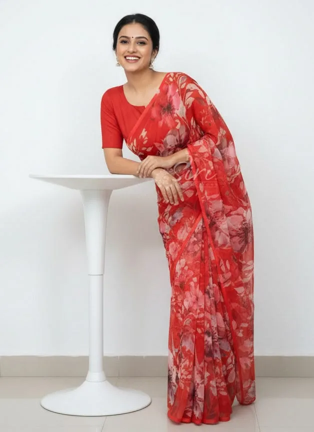 Woman wearing a red floral printed saree with a matching blouse, smiling while leaning on a white table. This monsoon-friendly saree features a lightweight sheer fabric with floral prints, making it a comfortable choice for festive occasions, casual outings, and everyday ethnic wear.