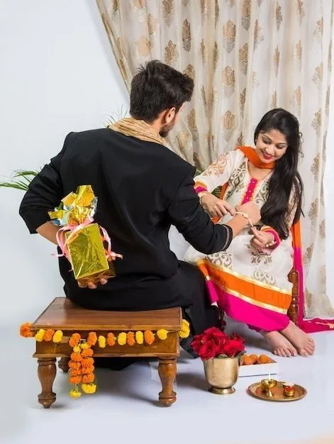 Raksha Bandhan 2026 Style Guide: Dress Up, Feel Good & Celebrate in Style 2 Raksha Bandhan 2026 Style Guide: Dress Up, Feel Good & Celebrate in Style A brother in a black kurta sits on a decorated wooden stool while his sister in a white and orange ethnic outfit ties a Rakhi on his wrist. Festive decorations, flowers, gifts, and a traditional puja thali surround them.