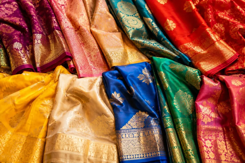 A vibrant collection of Indian saree fabrics in rich shades of purple, pink, red, gold, teal, yellow, ivory, blue, green, and magenta, featuring intricate floral motifs, paisley patterns, and ornate gold zari borders. Options to consider while shopping for a saree online