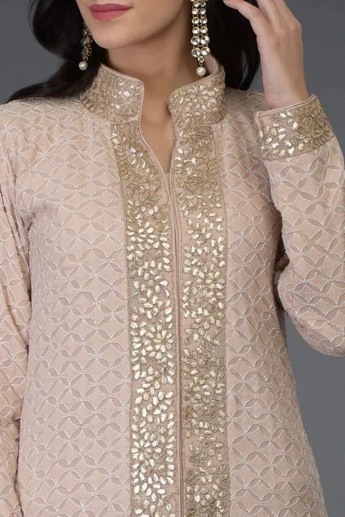 Elegant woman wearing a blush beige embroidered kurta with intricate gold floral embellishments and a patterned neckline and cuffs.
